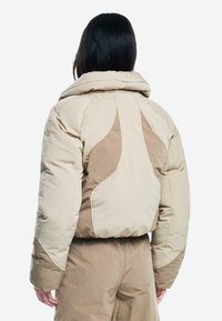Chaqueta de plumas beige con un alto collar, con acentos en tono marrón claro y costuras texturizadas, diseñada para ofrecer calor y comodidad.