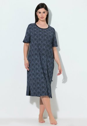 Vrouw met steil donker haar, gekleed in een knielange marineblauwe jurk met lichtblauwe sterrenpatronen, staat blootsvoets tegen een effen witte achtergrond.