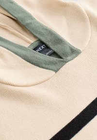 Beige neule vihreällä hupun reunalla, jossa on teksturoitu pinta ja musta vaaka-raita. Tägi näkyvissä kaula-aukossa.