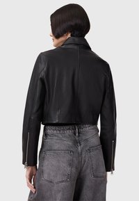 Chaqueta corta de cuero negra con cuello puntiagudo y puños con cremallera, combinada con pantalones gris de cintura alta con bolsillos traseros.