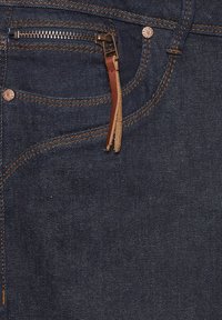 Jeans de mezclilla oscura con un bolsillo con cremallera y acentos de cuero. Cuenta con remaches de cobre y costuras naranjas en contraste. Tejido texturizado.