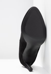 Chaussure à talon haut noire à bout pointu, présentée en angle pour montrer la semelle rainurée et le talon fin sur un fond blanc en coin.