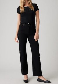 Schwarze hochtaillierte Jeans mit geradem Bein, aus Denim gefertigt. Kombiniert mit einem enganliegenden schwarzen Kurzarmoberteil und schwarzen Ballerinas. Glatte Textur.