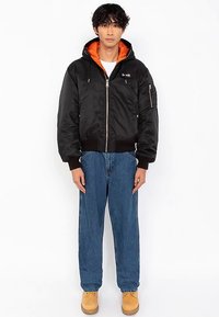 Chaqueta bomber negra con forro naranja, cierre de cremallera, puños y dobladillo acanalados, combinada con jeans azules y botas color marrón claro. Vista frontal en un modelo.