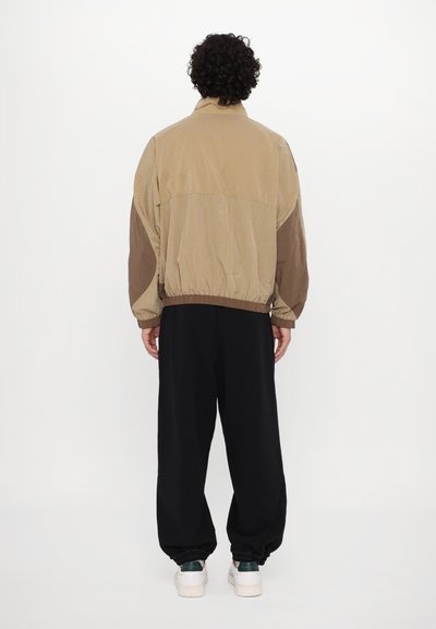 Jachetă în culori khaki și brună, cu guler înălțat, tiv elastic și croială largă, asortată cu pantaloni negri evazați și adidași.