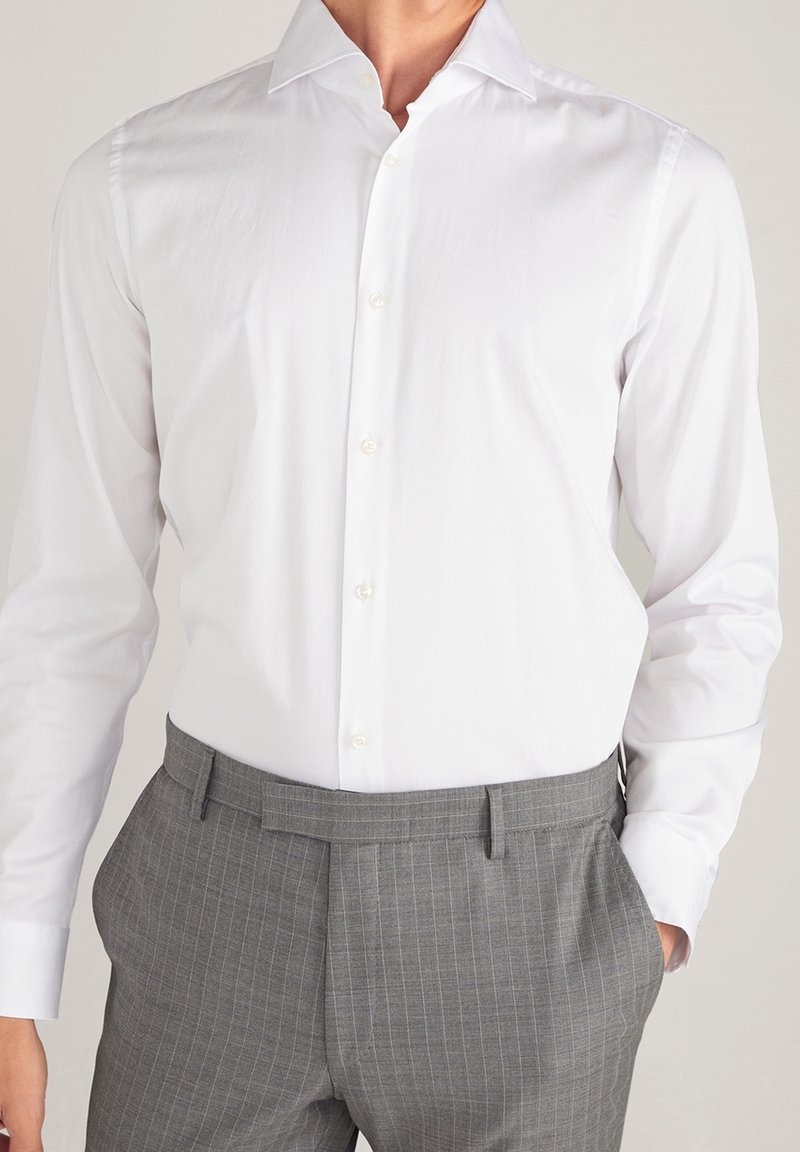 Camisa blanca de manga larga con cuello puntiagudo, cierre frontal de botones y textura lisa, combinada con pantalones grises con rayas finas.