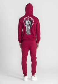 Bordový mikinový a teplákový set, s veľkým grafickým zobrazením anjela na chrbte, obklopeným bielym textom. Materiál vyzerá mäkko a štruktúrovane.