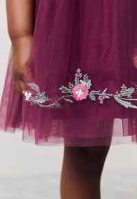 Vinröd tyllklänning med blommig broderi i rosa, grönt och vitt längs fållen, med en mjuk, lager-på-lager-textur och genomskinlig avslutning.