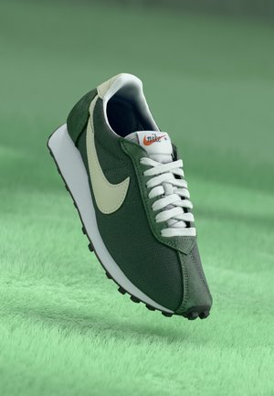 Zielony sneaker Nike z białym logo i sznurowadłami unoszący się nad zieloną, trawiastą powierzchnią.