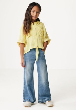 Camisa amarilla de manga corta abotonada con un lazo en la parte delantera, combinada con pantalones vaqueros de mezclilla azul de pierna ancha. Tejido suave y ajuste casual, con bolsillos laterales.