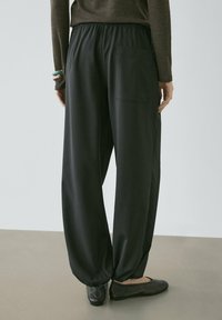 Person iført løstsiddende sorte bukser med elastiske ankler og en baglomme, kombineret med sorte flade sko og en brun langærmet top.