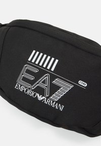 Juodas nailono juosmens krepšys su balta vertikalių juostelių dizainu ir "EA7 EMPORIO ARMANI" logotipu. Uždarymas užtrauktuku su reguliuojama juosta.