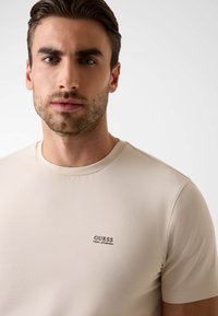 Beżowy prążkowany T-shirt z okrągłym dekoltem, z małym ciemnym logo "GUESS" z przodu. Wykonany z miękkiego, teksturowanego materiału.