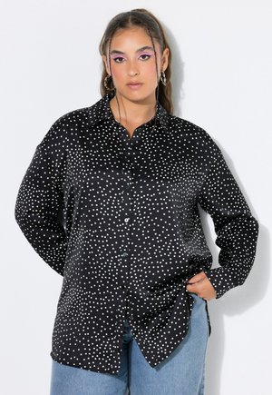 Kvinde med langt hår iført en sort skjorte med hvide polkaprikker og blå jeans, poserer med den ene hånd i lommen på hvid baggrund.