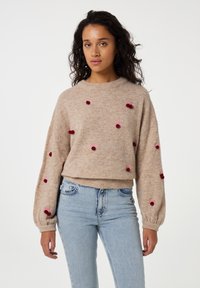 Beige Pullover mit langen, ballonförmigen Ärmeln und dekorativen roten Pompons. Weicher Stoff, runder Halsauschnitt, cropped Schnitt. Kombiniert mit hellblauen Jeans.