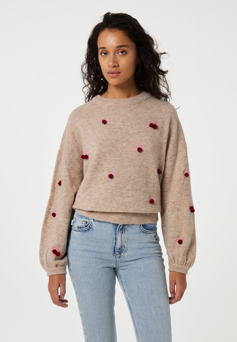 Beige Pullover mit langen, ballonförmigen Ärmeln und dekorativen roten Pompons. Weicher Stoff, runder Halsauschnitt, cropped Schnitt. Kombiniert mit hellblauen Jeans.