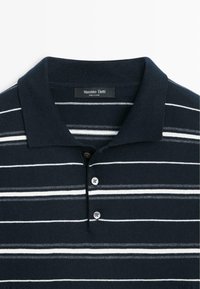 Marineblauw poloshirt met witte horizontale strepen, gemaakt van zachte stof. Voorzien van een kraag, een knoopsluiting met drie knopen en een label bij de nek.