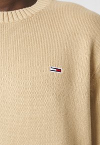 Pull en maille de couleur fauve avec un col côtelé et un petit logo de drapeau brodé en rouge, blanc et bleu sur la poitrine gauche.