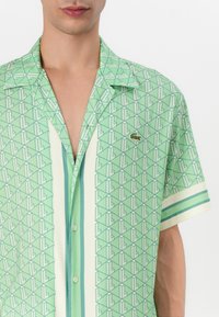 Short-sleeve green shirt with geometric pattern and cream vertical stripes. Features a small logo patch and lightweight fabric texture.