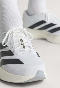 Weiße Laufschuhe mit schwarzen Streifen und "ADIZER0"-Text, getragen mit weißen strukturierten Socken auf einer hellen Oberfläche.