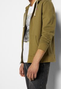 Olivgrön zip-up hoodie med kängurufickor, vita dragsnören och broderad logotyp. Mjukt tyg med ribbade ärmslut och nederkant.