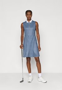 Zils rakstains golfa krekls ar apkakli, rāvējslēdzēju un plisētu svārku daļām, saskaņots ar baltām sporta kurpēm un golfa nūju.