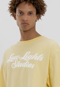 T-shirt en coton jaune avec le texte en cursive blanche "Low Lights Studios", coupe décontractée, col rond et manches courtes.
