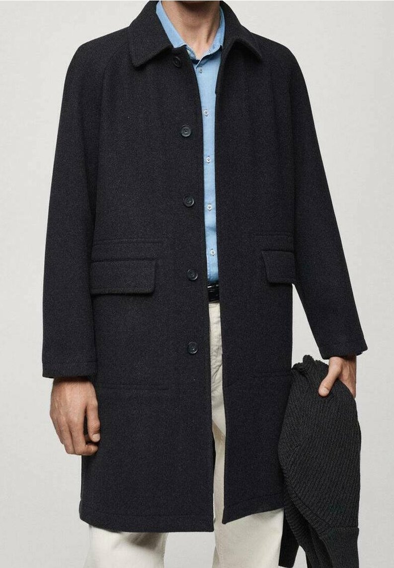 Homme portant un manteau sombre boutonné sur une chemise bleue et un pantalon clair, tenant dans une main un pull en maille foncé plié.