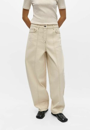 Mujer con pantalones beige de talle alto y pierna ancha con costuras visibles, camiseta beige de manga corta y zapatos negros con punta cuadrada.