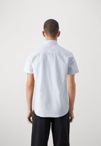 Camisa de manga corta azul claro con collar clásico, textura suave y dobladillo recto, capturada desde la parte posterior. Pantalones negros parcialmente visibles.