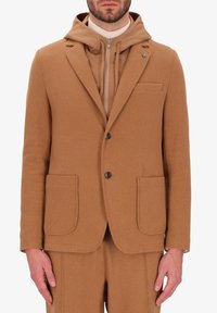 Blazer in lana color cuoio con rever a notched, due tasche frontali e chiusura con un solo bottone; indossato sopra un hoodie con zip coordinato in color cuoio.