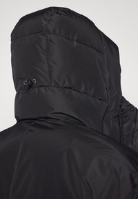 Giacca a vento nera con colletto alto e cappuccio. Presenta una superficie liscia e lucida e dettagli elastici per fissare il cappuccio.