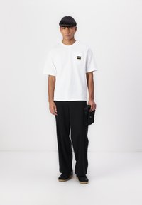 White short-sleeve t-shirt with a square pocket featuring a black label. Paired with black wide-leg trousers and black sneakers, textured fabric.