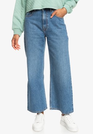 Jeans in denim a gamba larga di colore azzurro chiaro con vita alta. Presentano un classico design a cinque tasche e orlo grezzo. Indossati con sneakers bianche.