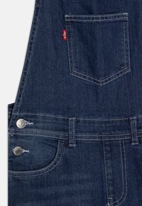 Džínové lacláče v tmavě modré barvě s kontrastním stehem. Mezi funkce patří nastavitelné popruhy, kovové doplňky a zadní kapsa.