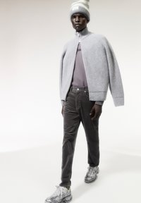 Model draagt een lichtgrijze ribgebreide vest met rits over een grijs t-shirt, gecombineerd met donkergrijze corduroypantalons en grijze sneakers met accenten.