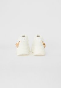 Baskets blanches avec une semelle épaisse, présentant un design texturé et un accent décoratif doré à l'arrière. Matériau lisse.