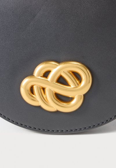 Μαύρη δερμάτινη τσάντα με ένα εμφανές χρυσό μεταλλικό σχέδιο κόμπου ως κλείσιμο, με λεπτή ραφή κατά μήκος των άκρων και λεία υφή.