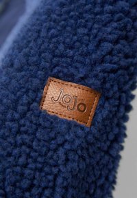 Temno modra frotirna tkanina z teksturirano, puhasto površino. Ima rjavo usnjeno oznako z vtisnjenim napisom "JoJo", prišito ob robu.
