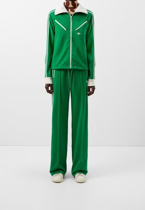 Persoon draagt een groen Adidas trainingspak met witte strepen, een jack met rits en diagonale zakken, wijduitlopende broek en witte sneakers.