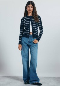 Námořnický pruhovaný cardigan s bílými pruhy, kulatým výstřihem a zapínáním na knoflíky, kombinovaný s džínami s vysokým pasem a rozšířenými nohavicemi a černými balerínami.