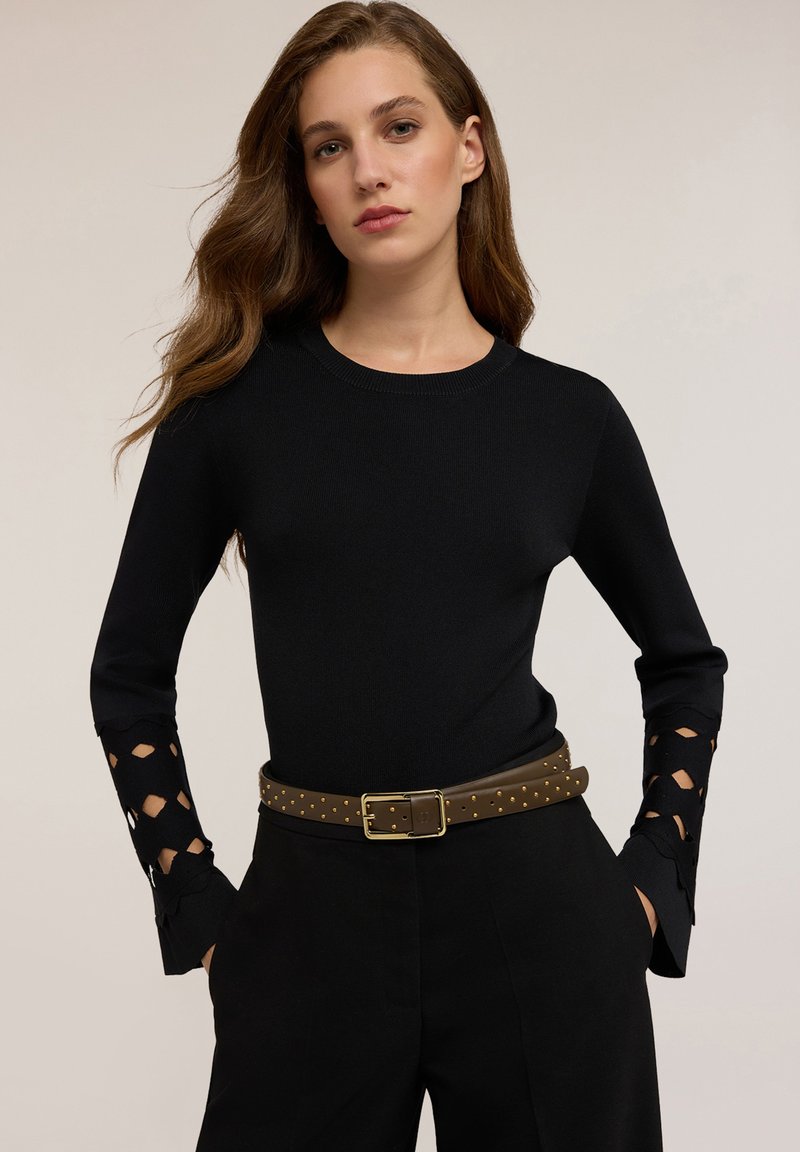 Femeie îmbrăcată cu un top negru cu mâneci lungi și cu detalii din dantelă, pantaloni negri și un curea maro cu ținte, stând cu mâinile în buzunare.