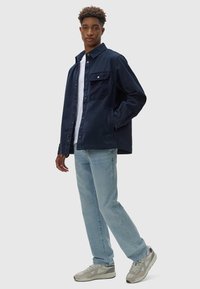 Giacca blu navy con bottoni, dotata di una tasca sul petto in corduroy, indossata sopra una maglietta bianca e jeans azzurri, abbinata a sneaker grigie.
