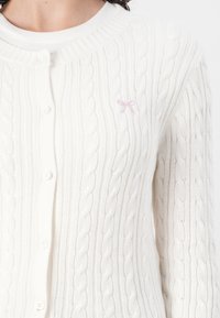 Hvit kabelstrikket cardigan med rund hals, knappelukking og en liten rosa sløyfe-logo på brystet. Strukturert stoff med ribbede detaljer.