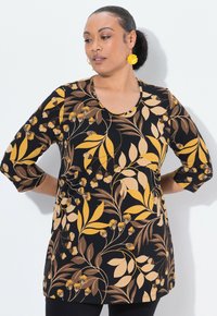 Vrouw met haar in een bun, draagt gouden bloemvormige oorbellen en een zwarte blouse met lange mouwen met een gele en bruine bloemenprint, handen achter de rug.