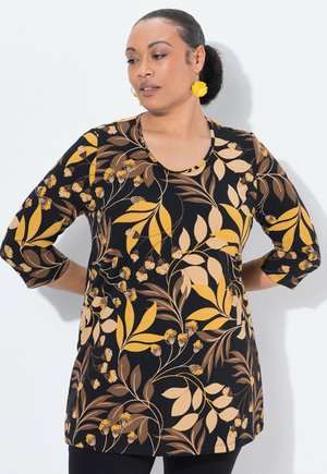 Frau mit einem Haarknoten, die goldene blumenförmige Ohrringe und ein schwarzes Langarmshirt mit gelbem und braunem Blumenmuster trägt, die Hände hinter dem Rücken.