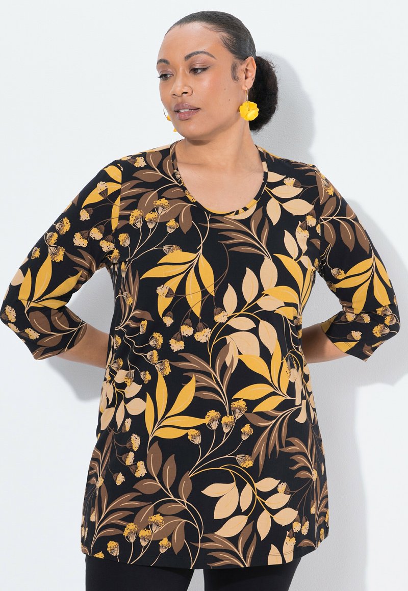 Vrouw met haar in een bun, draagt gouden bloemvormige oorbellen en een zwarte blouse met lange mouwen met een gele en bruine bloemenprint, handen achter de rug.
