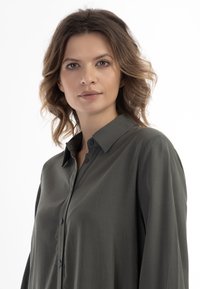 Camicia verde con bottoni, dal taglio comodo, dotata di colletto, maniche lunghe e texture liscia, esposta su uno sfondo bianco.