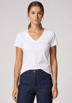 Femme aux cheveux bruns attachés, vêtue d'un t-shirt blanc uni à manches courtes avec col en V et d'un jean bleu foncé taille haute, debout devant un fond gris.
