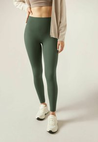 Green leggings made of stretchy fabric with a snug fit, paired with white sneakers and a beige cardigan. Smooth texture, high waist design.